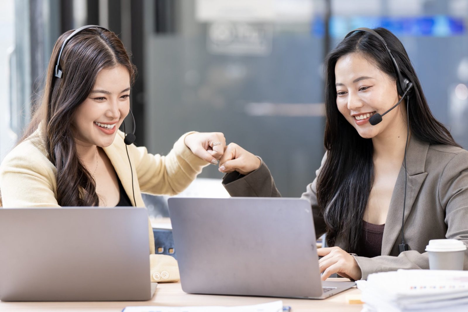 Two happy female customer service agents giving a fist bump while working with laptops and headsets, celebrating success in a modern office.